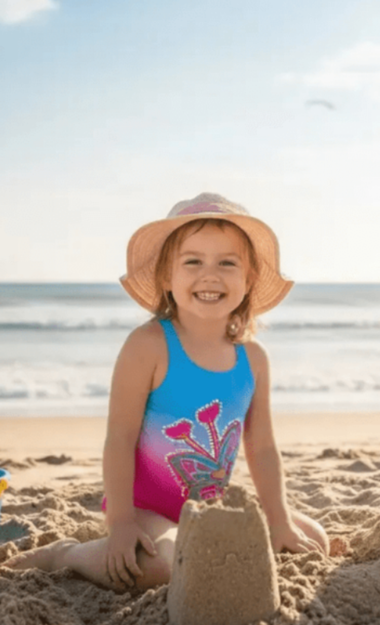 LuvBug girls one-piece swimsuit with pretty bug print, model on beach building sandcastle, made in Canada